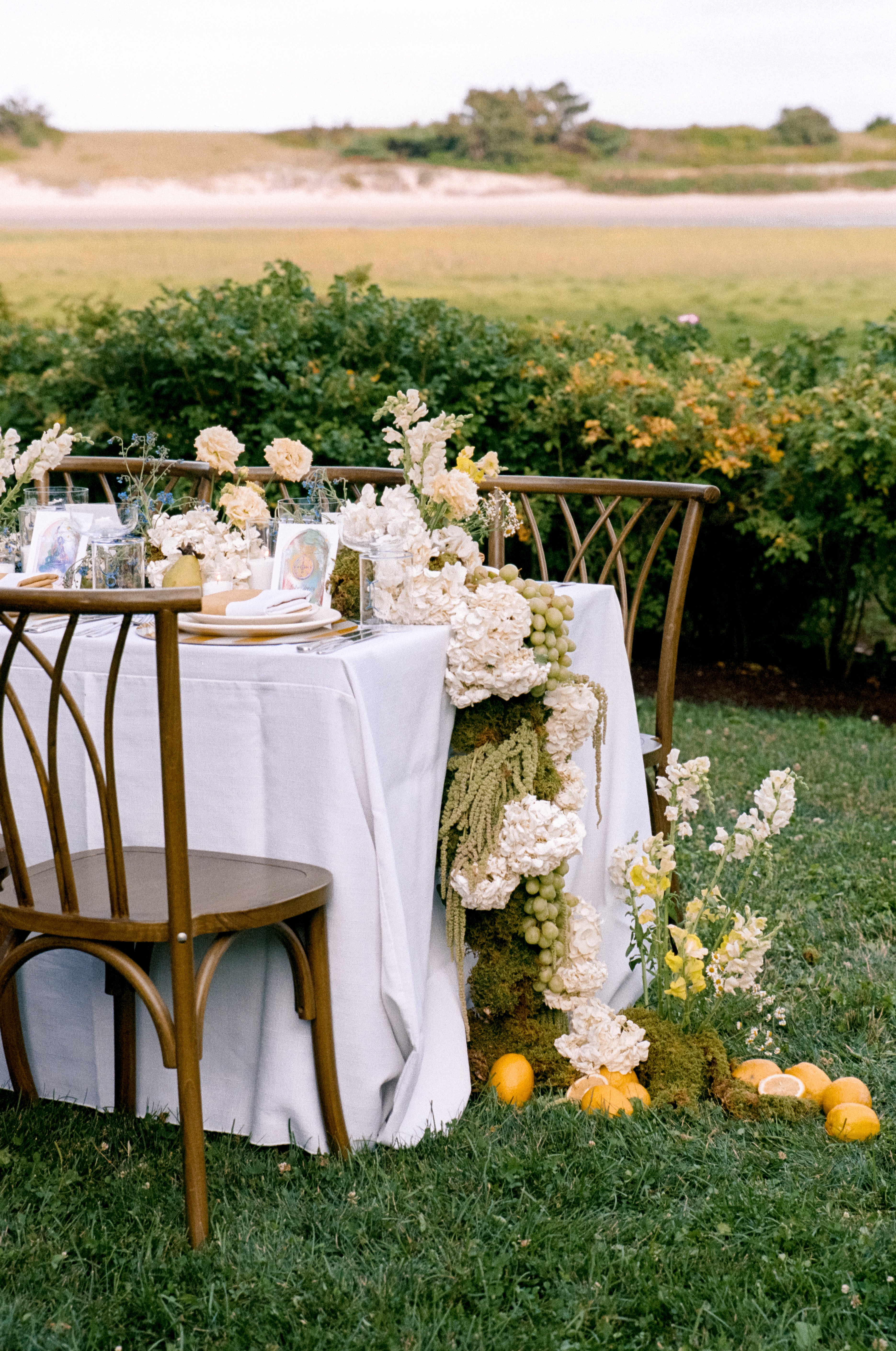 Elegant outdoor dining setup with white tablecloth, floral arrangements, and fruit on a grassy field.